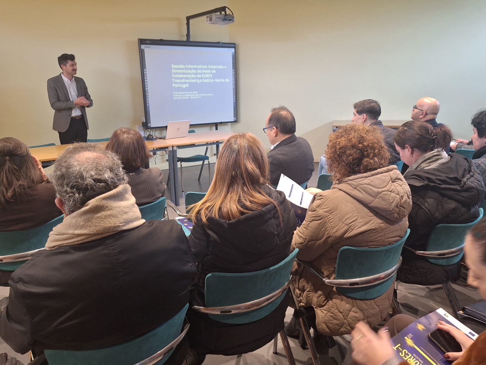 Pessoas sentadas num auditório com um dos palestrantes de pé junto a uma tela de projeção