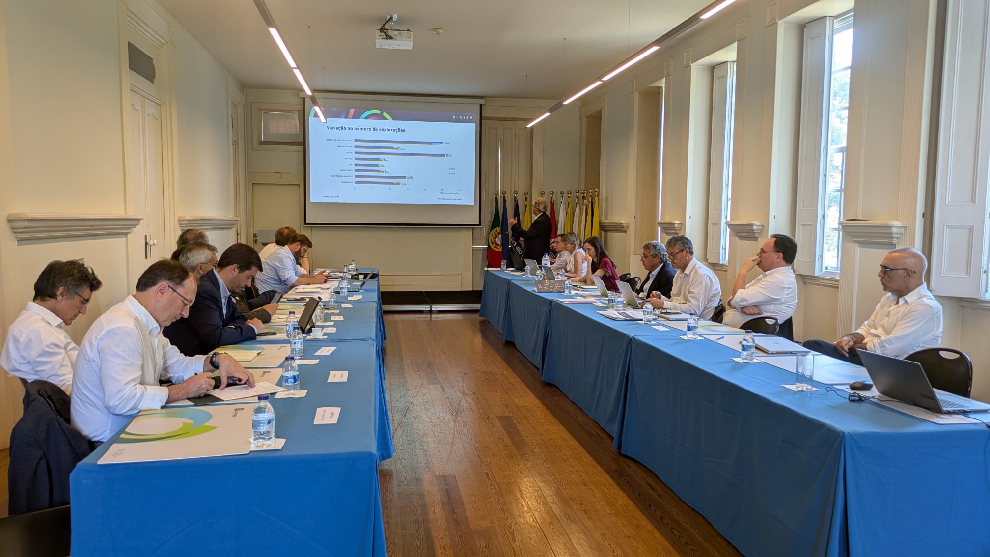 na imagem surgem dois corredores de mesas com os representantes do Alto Minho e da CCDRNORTE sentados de frente. Ao fundo da sala o Presidente da CCDRN de pé faz a sua intervenção com recurso a uma projeção.